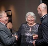 Three people in formal attire are laughing and holding champagne glasses at an indoor event.