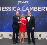 Three people in formal wear on stage hold a trophy. The background displays "Winner Jessica Lambert" with abstract blue graphics.