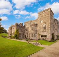 A historic stone castle with towers stands against a blue sky. It's surrounded by a green lawn and a walking path.