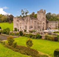 A large stone mansion with multiple towers sits amid expansive green lawns and gardens, surrounded by trees under a blue sky.
