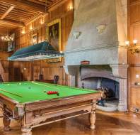 A billiards table in a wood-paneled room with a large stone fireplace, paintings, and a staircase in the background.