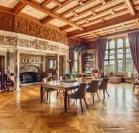 A spacious, ornate room with a large fireplace, wooden ceiling, bookshelves, and seating area. A table with chairs is on the wooden floor. Large windows allow natural light.