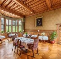 Elegant dining room with wood paneling, large windows, and round tables set with white tablecloths and chairs. A portrait hangs on the wall above decorated side tables and potted plants.