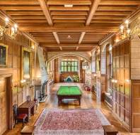 A grand room with wooden paneling features a billiard table, chandeliers, a large rug, and a stone fireplace. Walls are adorned with paintings and windows provide natural light.