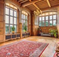 Grand room with large windows, wood-paneled walls, a red patterned rug, chandeliers, and furniture, including a sofa and chairs. Green plants by the windows.
