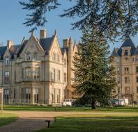 A large historic building with ornate architecture is surrounded by a grassy area, trees, and a clear blue sky.