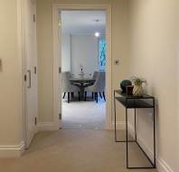 Hallway view of a modern, carpeted interior leading to a dining area with a round table and chairs. A small black console table with decorative items is on the right side.