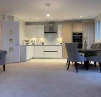 Modern open-plan living area with a kitchen, round dining table, and gray armchair. Neutral colors, wall oven, and decorative elements create a cozy ambiance.