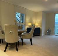 A minimalist dining room with a round wooden table, four beige upholstered chairs, a sideboard with two lamps, and a large window with a view.