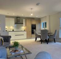 Modern open-plan kitchen and living area with white cabinets, wooden accents, dining table with chairs, and a sofa. Natural light enters through large windows.