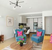 Modern living room with two blue armchairs, a yellow sofa, and a wooden coffee table. Bright pillows and plants add color. A kitchen is visible in the background, and a sleek light fixture hangs overhead.