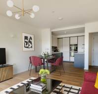 Modern living room with wood flooring, featuring a TV, coffee table, and vibrant colored furniture. Dining area with red chairs in the background, leading to a minimalist kitchen.