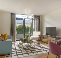 Modern living room with a blue sofa, glass coffee table, and TV. Pink dining chairs and a geometric rug are featured, with large windows overlooking a balcony with greenery.