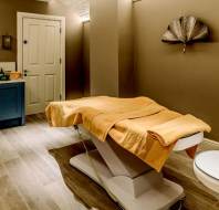 A massage room with a treatment table, covered with a yellow towel, a sink with toiletries, and decorative wall fans.