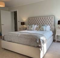 Bedroom with a large upholstered bed, pillows, and a blanket. Two bedside tables with lamps are on each side. A green chair is in the corner. The room has beige walls and a carpeted floor.