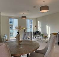 Bright living and dining area with modern decor, featuring a round table, gray upholstered chairs, a sofa, armchairs, and large windows with views of adjacent buildings.