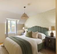 A cozy bedroom with a large bed, green velvet headboard, two bedside tables with lamps, a textured wall, and a window with partially open curtains allowing natural light in.