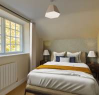 A cozy bedroom with a bed featuring white and mustard bedding, two lamps on wooden side tables, and a window with beige curtains letting in natural light.