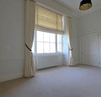 Empty room with beige carpet, large window with curtains, and a closed door. Walls are white and there's a black lampshade on the ceiling.
