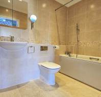 Modern bathroom with beige tiles, wall-mounted sink, toilet, and bathtub with a shower. A round mirror and ladder-style towel radiator are also visible.