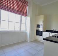 Bright kitchen with large windows, red checkered curtains, white cabinets, built-in oven and microwave, black countertops, and tiled floor.