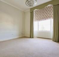 Empty room with beige carpet, white walls, and a large window with floral Roman shade and green curtains. Ceiling has a circular light fixture.