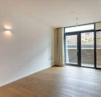 Empty room with wooden flooring, a single wall light, and large glass doors leading to a balcony. Simple, neutral design with curtains drawn to the sides.