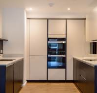 Modern kitchen with integrated appliances, dual countertops, and wood flooring, featuring minimalistic white and dark cabinetry.