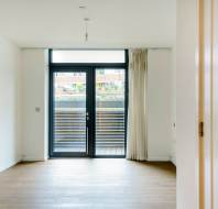Empty room with wooden floors, large window, and glass door leading to a small balcony. White walls and ceiling with a single light fixture.