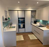 Modern kitchen with white cabinets, turquoise backsplash, wooden floor, and integrated appliances. Countertops hold a kettle, toaster, and other kitchen items. Wall shelves display decor.
