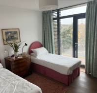 Bedroom with two single beds, wooden chest with flowers, a painting on the wall, and large window with curtains leading to a balcony.