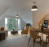 Modern attic apartment with a living area, dining table, and kitchen. Features sloped ceilings, large windows, and yellow chairs.
