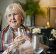 Older woman holding a glass, smiling while sitting indoors. She wears a colorful shawl; multiple wine glasses are in front. Warm lighting and a lamp in the background.