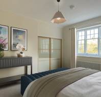 A bedroom with a bed, a console table with flowers and decorative items, two paintings on the wall, and a window with curtains.