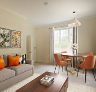 A modern living room with a gray sofa and orange pillows, floral wall art, a round dining table with orange chairs, and a pendant light above. Neutral tones throughout.
