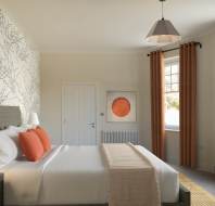 A bedroom with a bed featuring white linens and orange pillows. There are orange curtains, a patterned accent wall, a painting, and two windows letting in natural light.