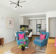Modern living room with blue and yellow chairs, a wooden coffee table, and a wall-mounted TV. A kitchen is visible in the background, and bright cushions add color to the space.
