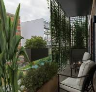 A small urban balcony with a chair, side table, and plants. Vertical trellis with greenery partially obstructs the view of neighboring buildings.