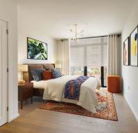 Modern bedroom with a large bed, colorful pillows, and a patterned rug. Two framed artworks hang on the white walls, and a window with sheer curtains offers a city view.