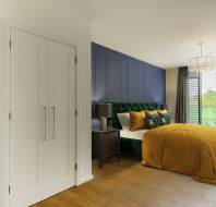 Modern bedroom with a large bed, mustard duvet, green upholstered headboard, and wooden flooring. There are white closets, a navy accent wall, and a window with a view of greenery.