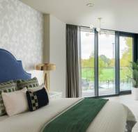 A modern bedroom with a blue headboard, patterned pillows, a brass lamp, and a green throw on the bed. Large windows show a view of trees and a sliding glass door leads to a balcony.