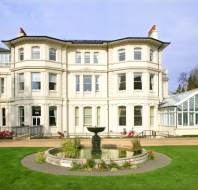 Large white Victorian-style building with multiple windows, a glass conservatory on one side, and a central fountain in a landscaped garden foreground.