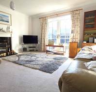 A cozy living room with a fireplace, TV, and bookshelves. There's a patterned rug, two armchairs, and a couch with cushions. Large window with curtains lets in natural light.