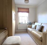 Small, tidy living room with a beige sofa, wooden cabinet, window with a fabric blind, and light-colored walls. A mirror rests on the cabinet, and two cushions are on the sofa.