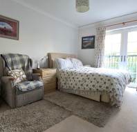 Bedroom with a double bed, plaid armchair, wooden nightstand, and large window with curtains. Walls are white with landscape paintings. Plush rug on beige carpeted floor.