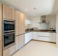 Modern kitchen with wooden cabinetry, double built-in ovens, a stovetop with a hood, and light-colored flooring. A dining room is visible through an open door to the right.