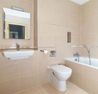 Modern bathroom with beige tiles, wall-mounted sink, mirror, toilet, and white bathtub with a handheld showerhead. Chrome towel rack visible on the left wall.