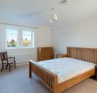 A minimalist bedroom with a wooden bed, white mattress, two small wooden drawers, a tall chest of drawers, a wooden chair, and a window with a view of greenery.