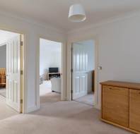A hallway with beige carpet leads to rooms with white doors. A wooden sideboard is on the right, and a hanging light fixture is above. Rooms include a kitchen and a living room with a sofa.