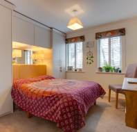 Bedroom with double bed covered in a patterned purple and red duvet. There are built-in wardrobes, a desk with a chair, two windows with blinds, and a small shelf with plants.